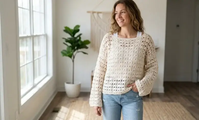 Woman wearing an off-white lightweight boho lace open stitch oversized crochet pullover over a tank top in a bright airy interior
