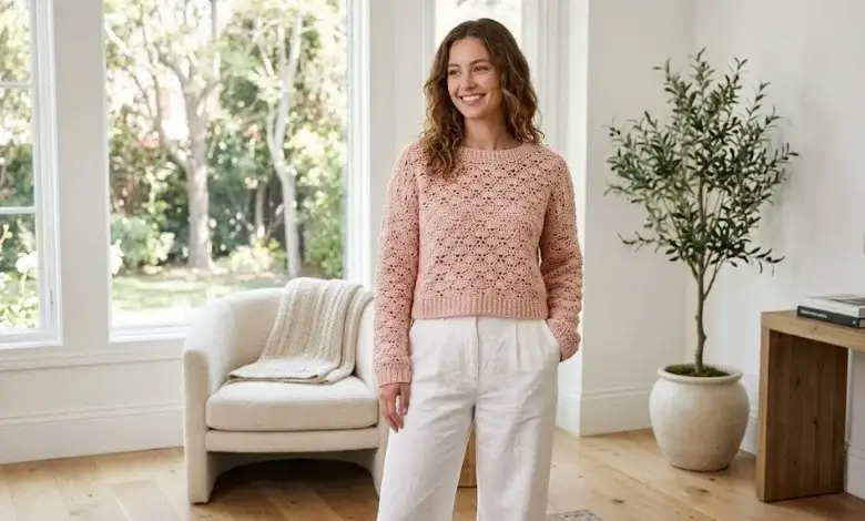 Woman wearing a blush pink cropped lace shell stitch crochet sweater with white linen trousers in a bright modern American home