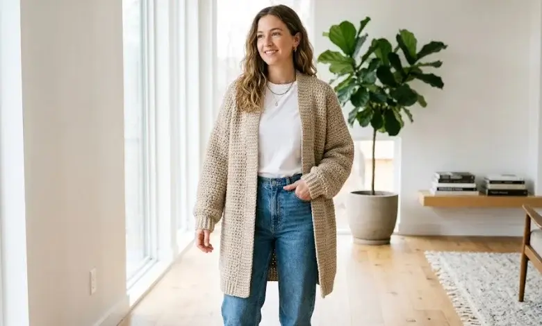 Woman wearing a long oatmeal beige oversized moss stitch crochet cardigan open front over a white tee with jeans in a minimalist interior