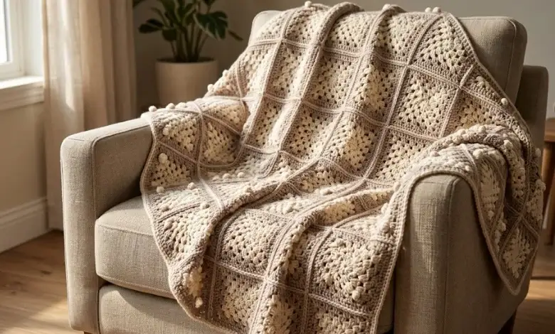 Taupe and cream granny square crochet blanket with raised bobble stitch texture draped over a neutral linen armchair in warm natural light