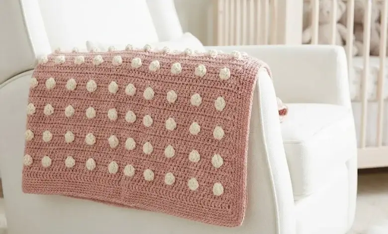 Cream and dusty rose baby crochet blanket with raised three-dimensional bobble stitch texture draped over a white nursing chair