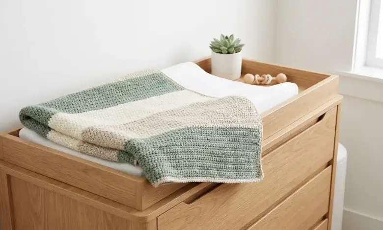 Modern baby crochet blanket with wide sage green cream and oatmeal stripe pattern folded on a light oak changing table