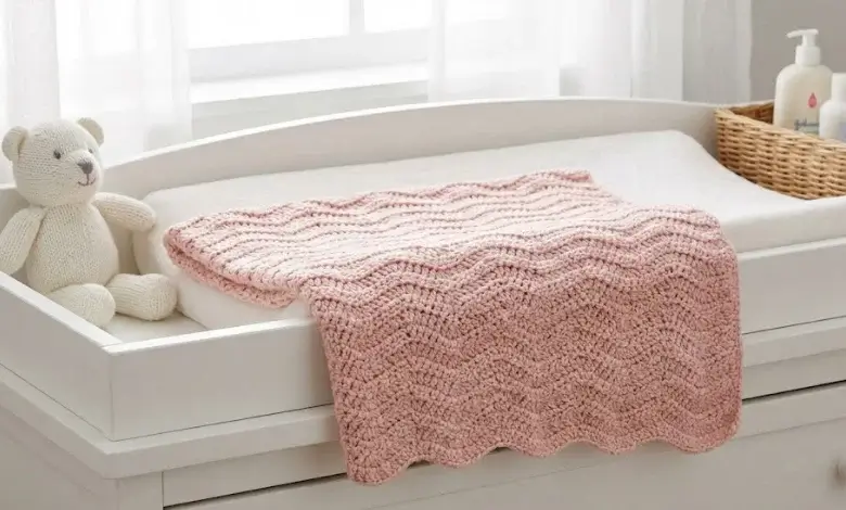 Blush pink crochet ripple stitch receiving blanket folded next to a white teddy bear on a changing table