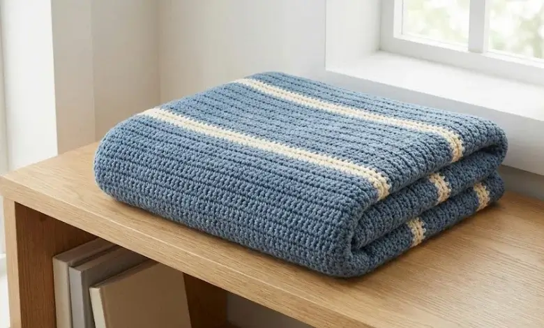 Minimalist crochet throw blanket with wide dusty blue color blocks and thin cream contrast stripes folded on a light oak shelf