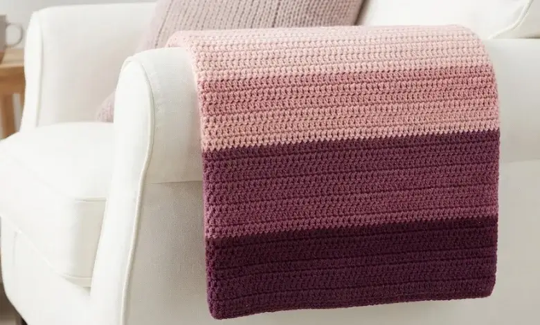 Gradient ombre crochet lap blanket transitioning from pale pink to deep plum in five stripe shades draped over a white armchair